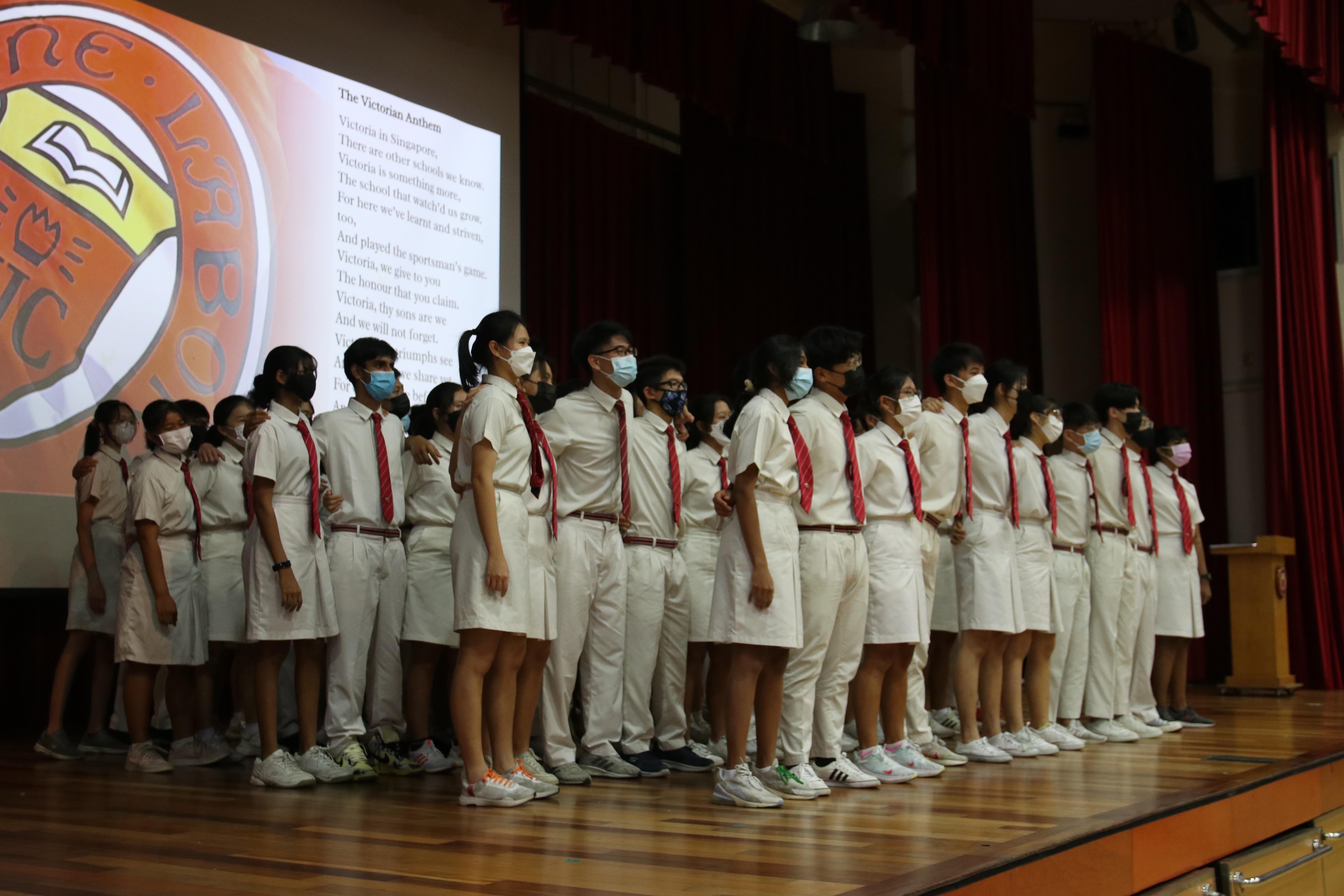 Students in white uniforms with red ties and face masks stand in a row on a school stage during an anthem ceremony.